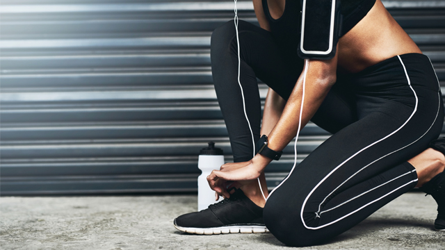 Woman in sport attire lacing up her running shoes before starting to run. She has a mobile phone with headphones, a smartwatch and a bottle of water.