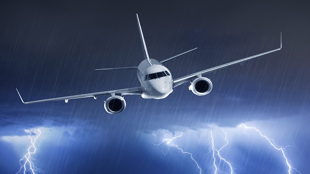 A passenger jet is flying out of dark clouds in a rainstorm with cloud-to-ground lightning strikes visible in lighter skies below.