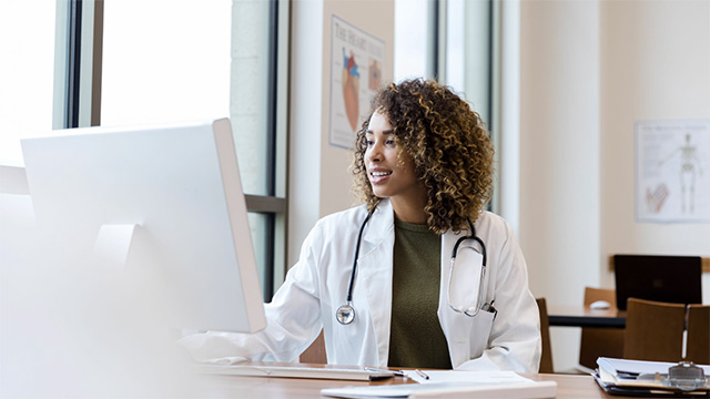 Female physician of color sitting at a desk staring at a monitor with a stethoscope around her neck. 