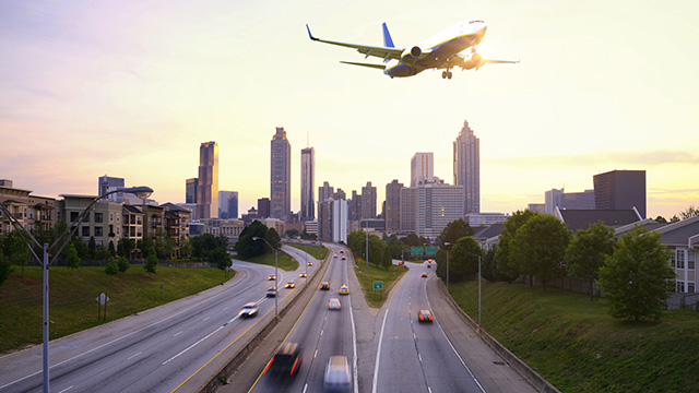 An airplane flies over several lanes of a highway, between treelined areas of residential buildings, with a city skyline in the background