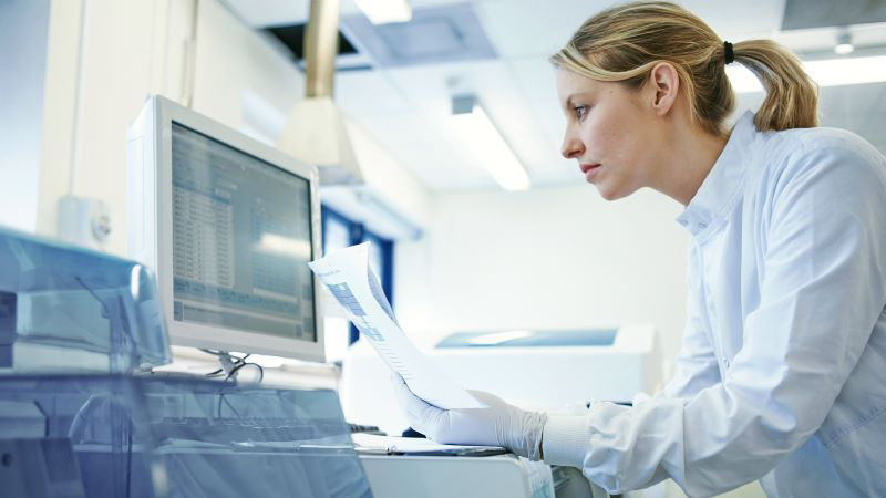 White female professional in a lab is using an integrated labeling system on a computer screen and reviewing a document.