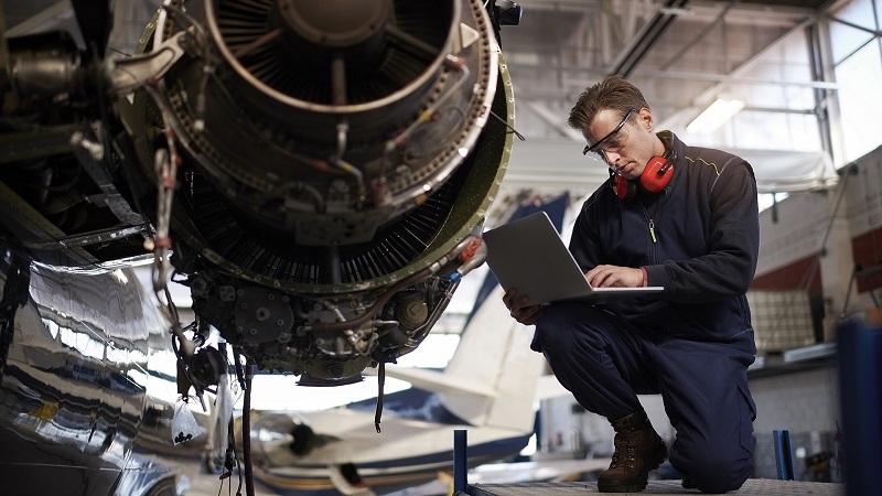 Aircraft engineer holding laptop computer while repairing and maintaining airplane jet engine.
